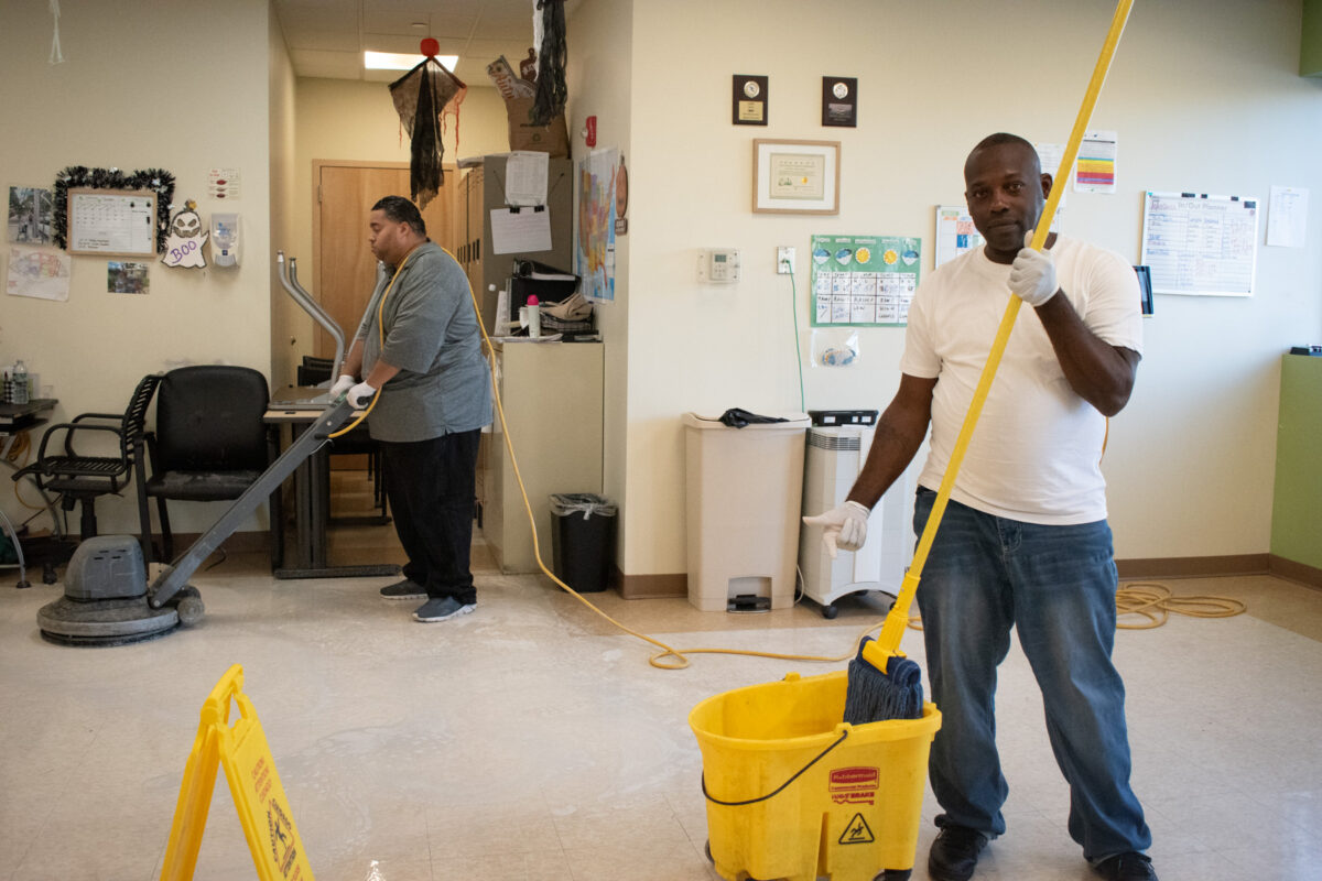 Salam Edmonds holds a mop and poses for a photo, while Louis Valentin practices using the buffer, in the Dorothy & Michael Styler Day Services Center of AHRC NYC, New York City, Oct. 5, 2023. (Credit: Angelica Ang)