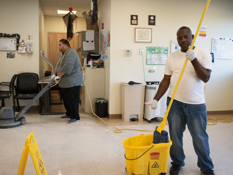 Salam Edmonds holds a mop and poses for a photo, while Louis Valentin practices using the buffer, in the Dorothy & Michael Styler Day Services Center of AHRC NYC, New York City, Oct. 5, 2023. (Credit: Angelica Ang)