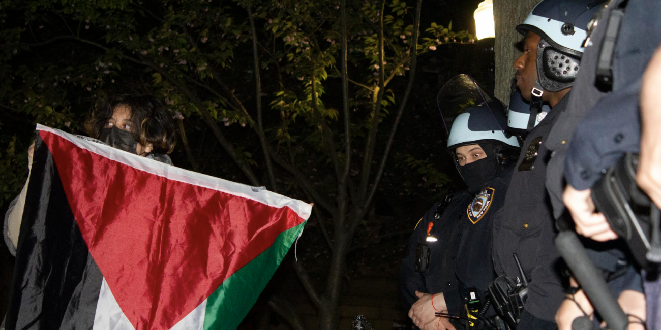 A protester holds the Palestinian flag aloft in front of NYPD officials on Tuesday, April 30, 2024 inside the Columbia University campus (Credit: Uzma Afreen)