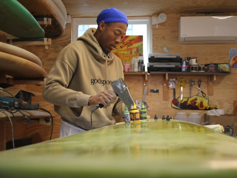 In his shipping-container workspace, Rockaway shaper Paul Godette repairs a dent in a client’s board, part of his effort to keep surfboards in use instead of tossed out. (Credit: Kate Waxman)