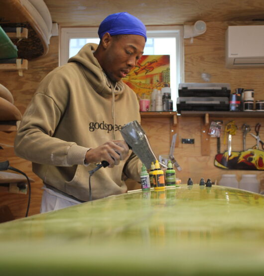 In his shipping-container workspace, Rockaway shaper Paul Godette repairs a dent in a client’s board, part of his effort to keep surfboards in use instead of tossed out. (Credit: Kate Waxman)